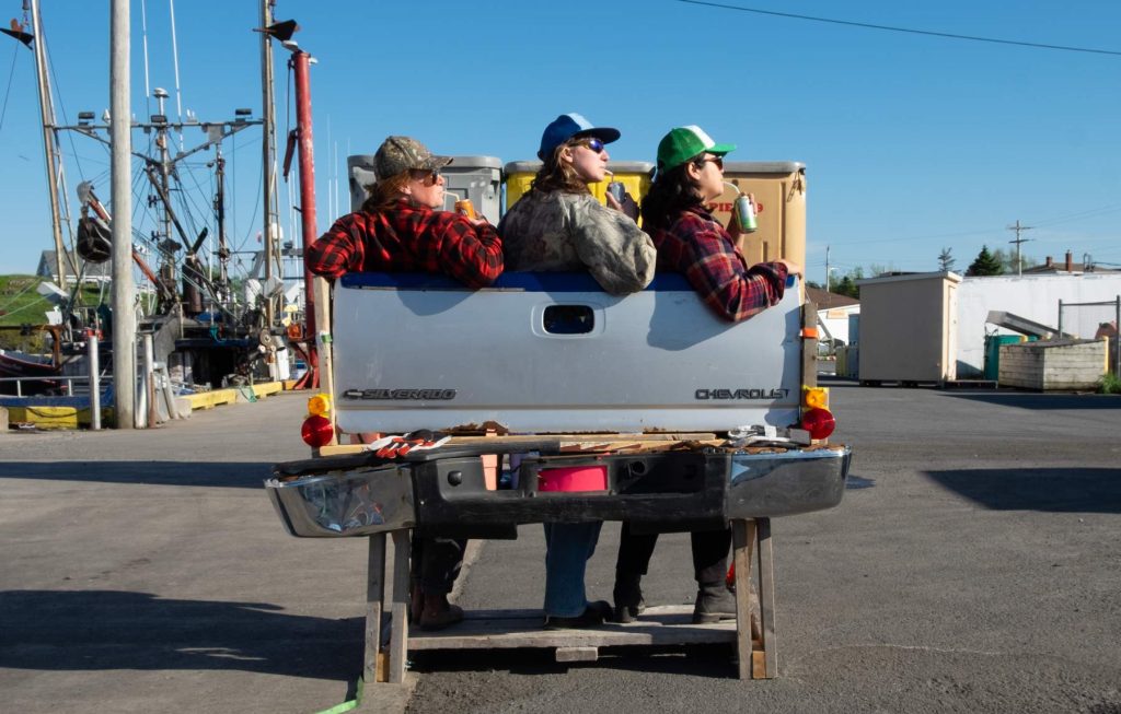 Artists Emily Neufeld, Drew Pardy, and Annie Canto in trucker hats lean against a freestanding tailgate sculpture looking in the same direction sipping on soda cans with bendy straws.